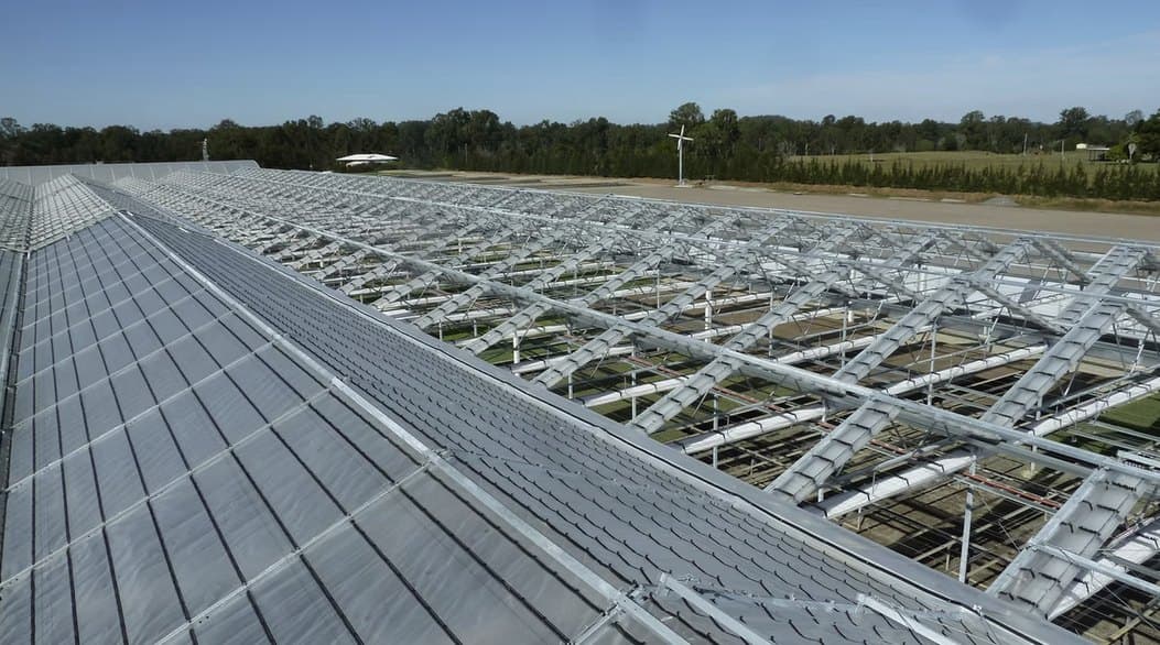 Exterior view across the steel roof structure of a large commercial greenhouse under a clear sky.